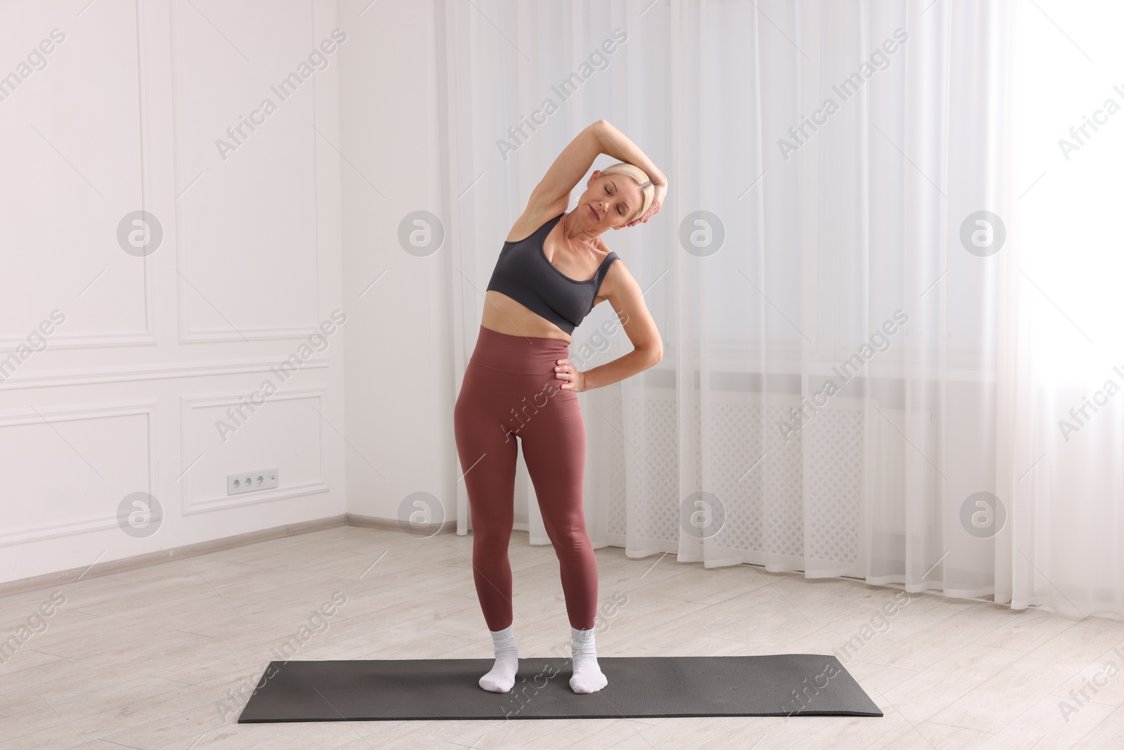Photo of Mature woman in sportswear stretching on mat near window at home