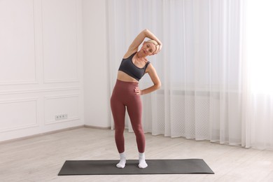 Photo of Mature woman in sportswear stretching on mat near window at home