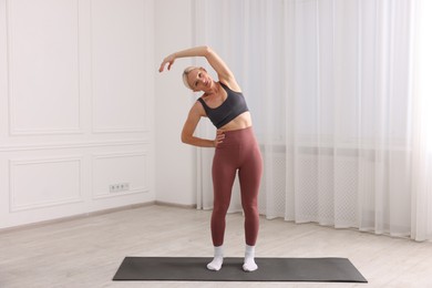 Photo of Mature woman in sportswear stretching on mat near window at home