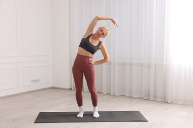 Photo of Mature woman in sportswear stretching on mat near window at home