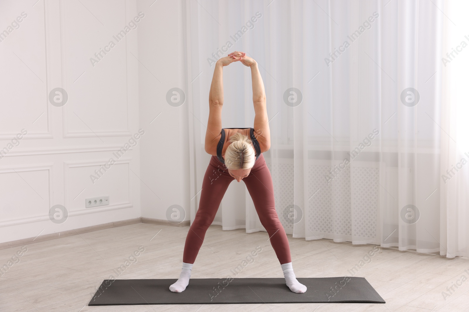 Photo of Mature woman in sportswear stretching on mat near window at home