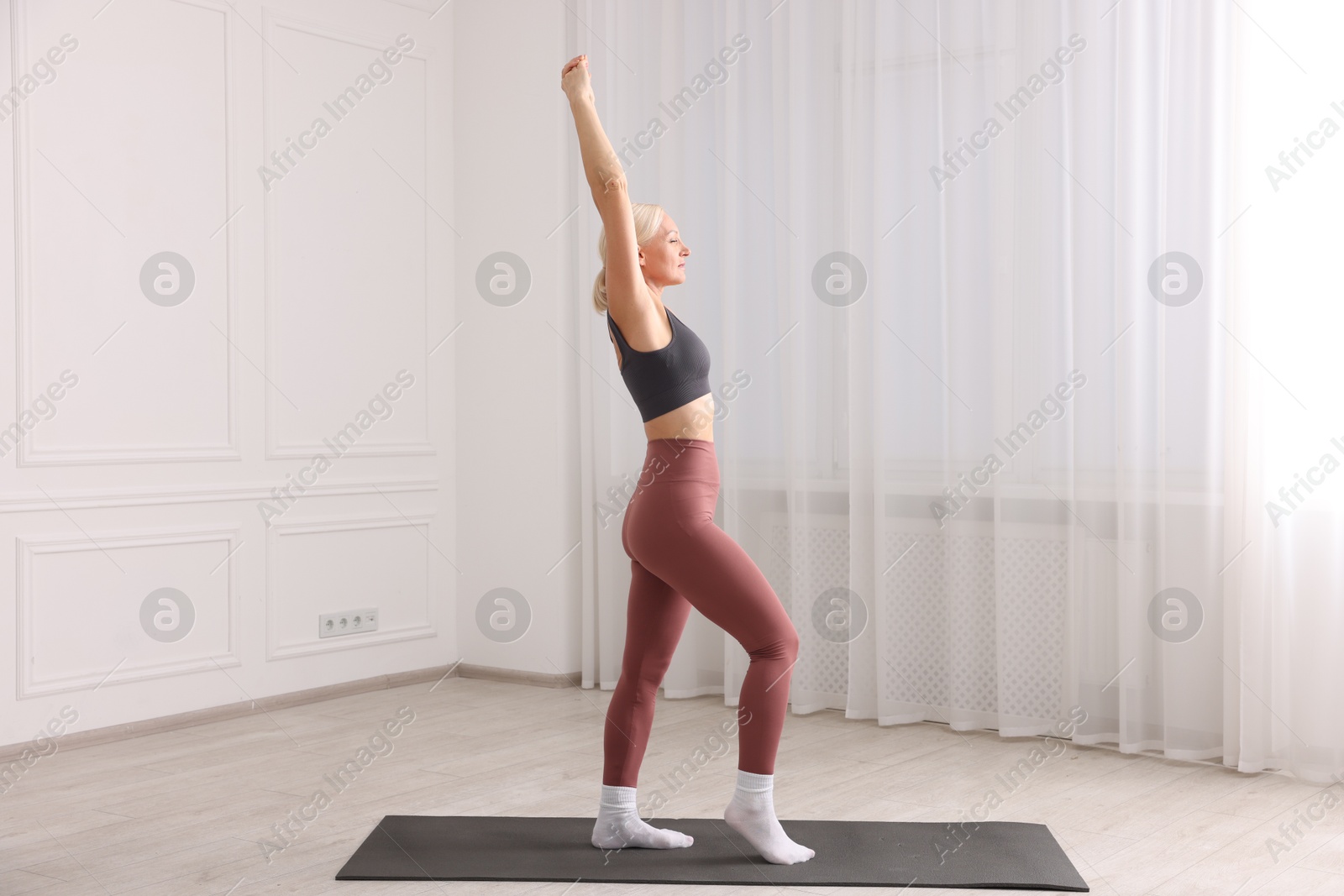 Photo of Mature woman in sportswear stretching on mat near window at home