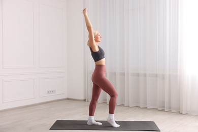 Photo of Mature woman in sportswear stretching on mat near window at home
