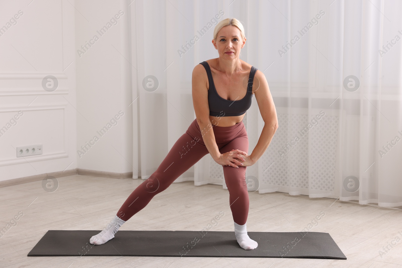 Photo of Mature woman in sportswear stretching on mat near window at home