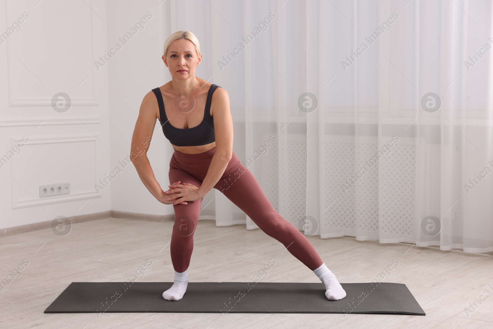 Photo of Mature woman in sportswear stretching on mat near window at home