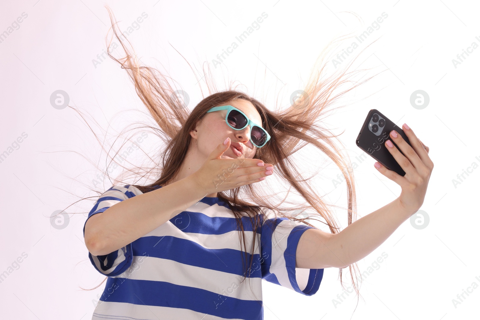 Photo of Happy woman in sunglasses with beautiful long hair using smartphone and sending air kiss on white background, low angle view