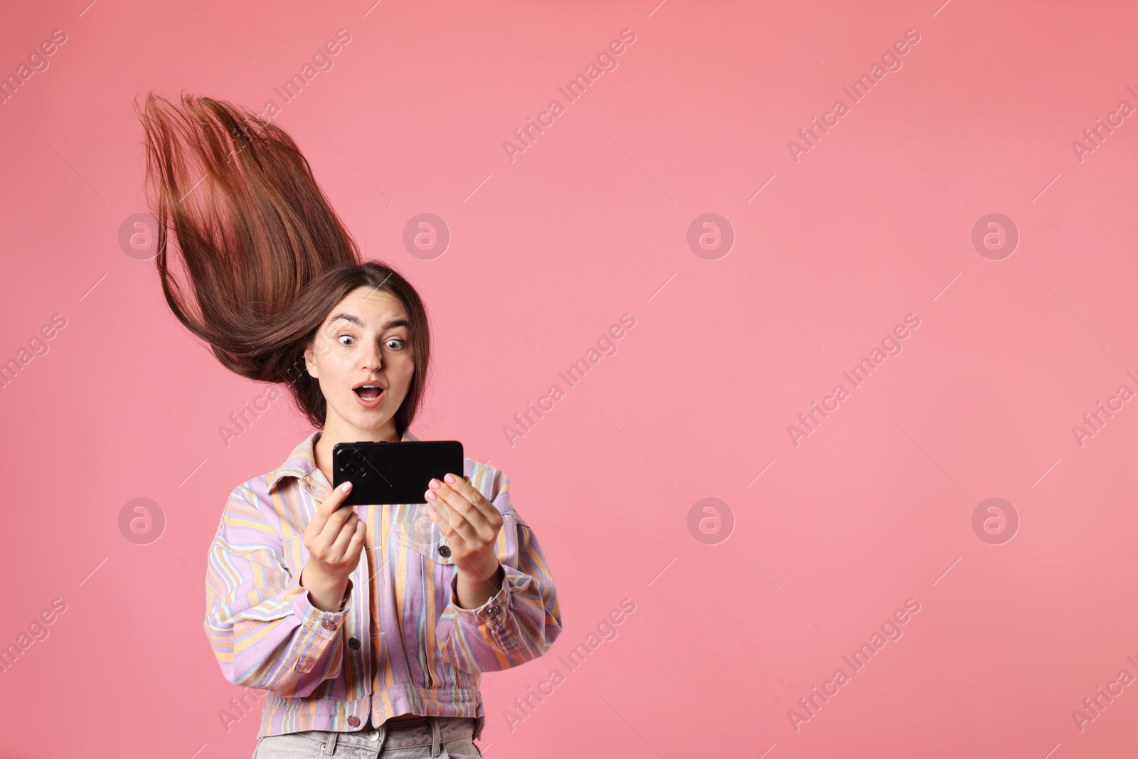 Photo of Surprised woman in jacket with beautiful long hair using smartphone on pink background. Space for text