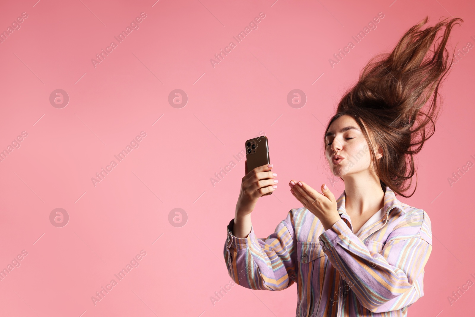 Photo of Young woman in jacket with beautiful long hair having videochat by smartphone and sending air kiss on pink background. Space for text