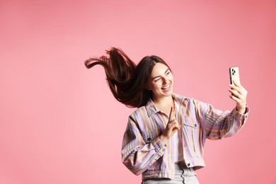 Photo of Happy woman in jacket with beautiful long hair taking selfie by smartphone and showing peace sign on pink background. Space for text