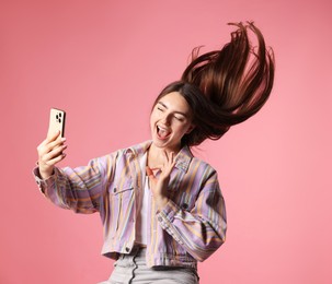 Photo of Happy woman in jacket with beautiful long hair taking selfie by smartphone and showing ok gesture on pink background
