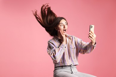 Photo of Surprised woman in jacket with beautiful long hair using smartphone on pink background. Space for text