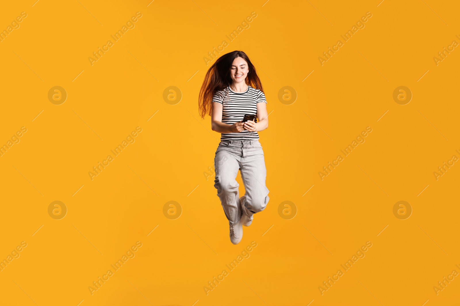 Photo of Happy woman with beautiful long hair using smartphone and jumping on orange background