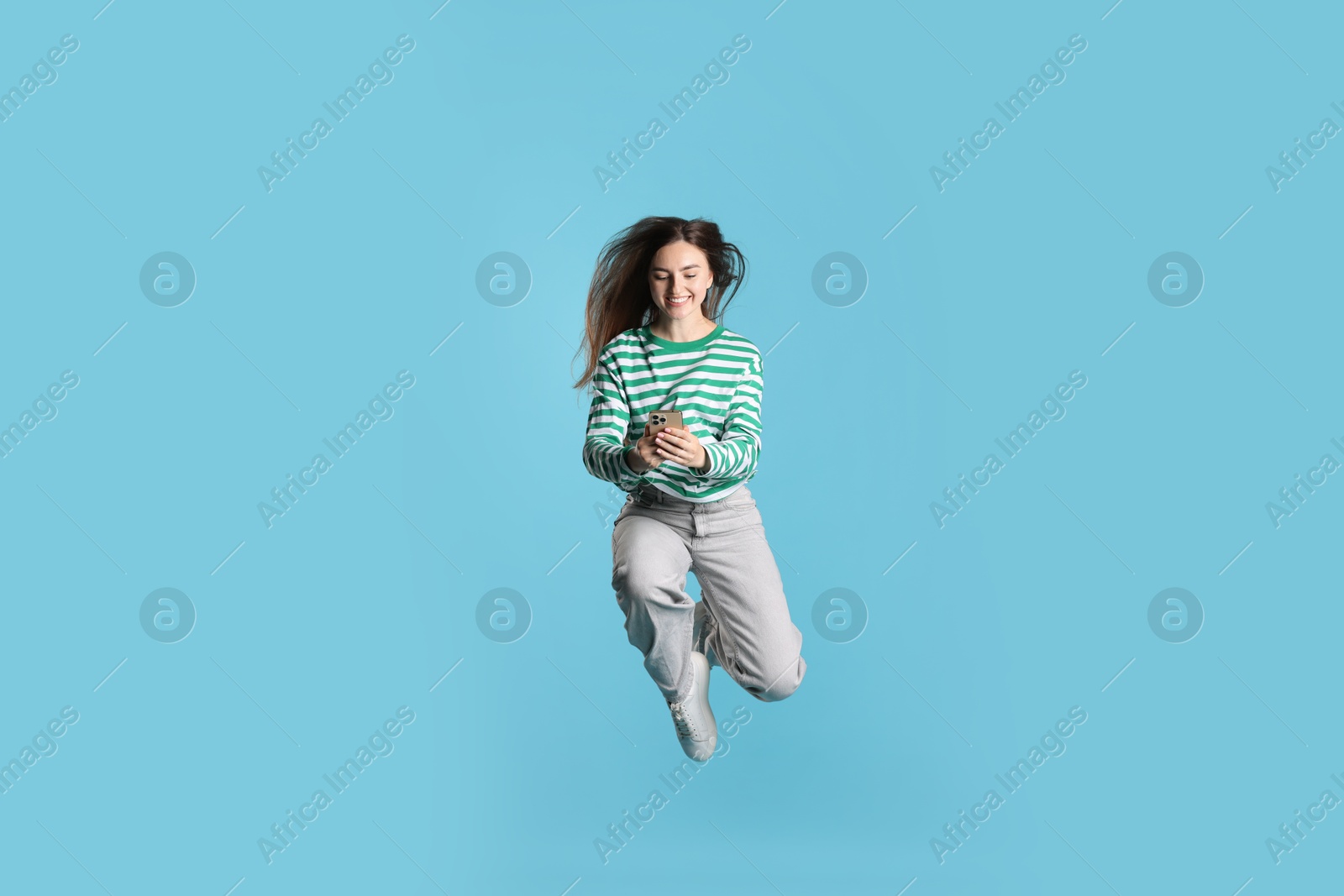 Happy woman with beautiful long hair using smartphone and jumping on light blue background Photo of Happy woman with beautiful long hair using smartphone and jumping on light blue background