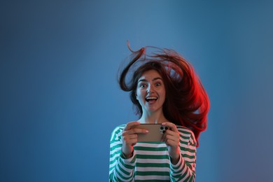 Photo of Happy woman with beautiful long hair using smartphone in neon lights against blue background. Space for text