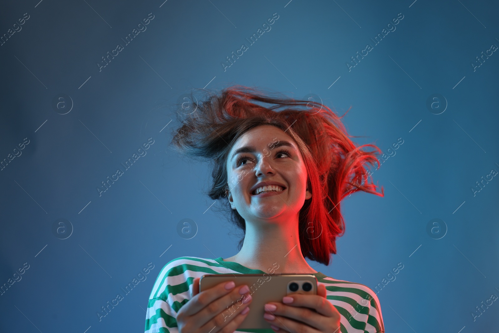 Photo of Happy woman with beautiful long hair using smartphone in neon lights against blue background, low angle view
