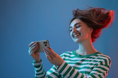 Photo of Happy woman with beautiful long hair using smartphone in neon lights against blue background, low angle view. Space for text