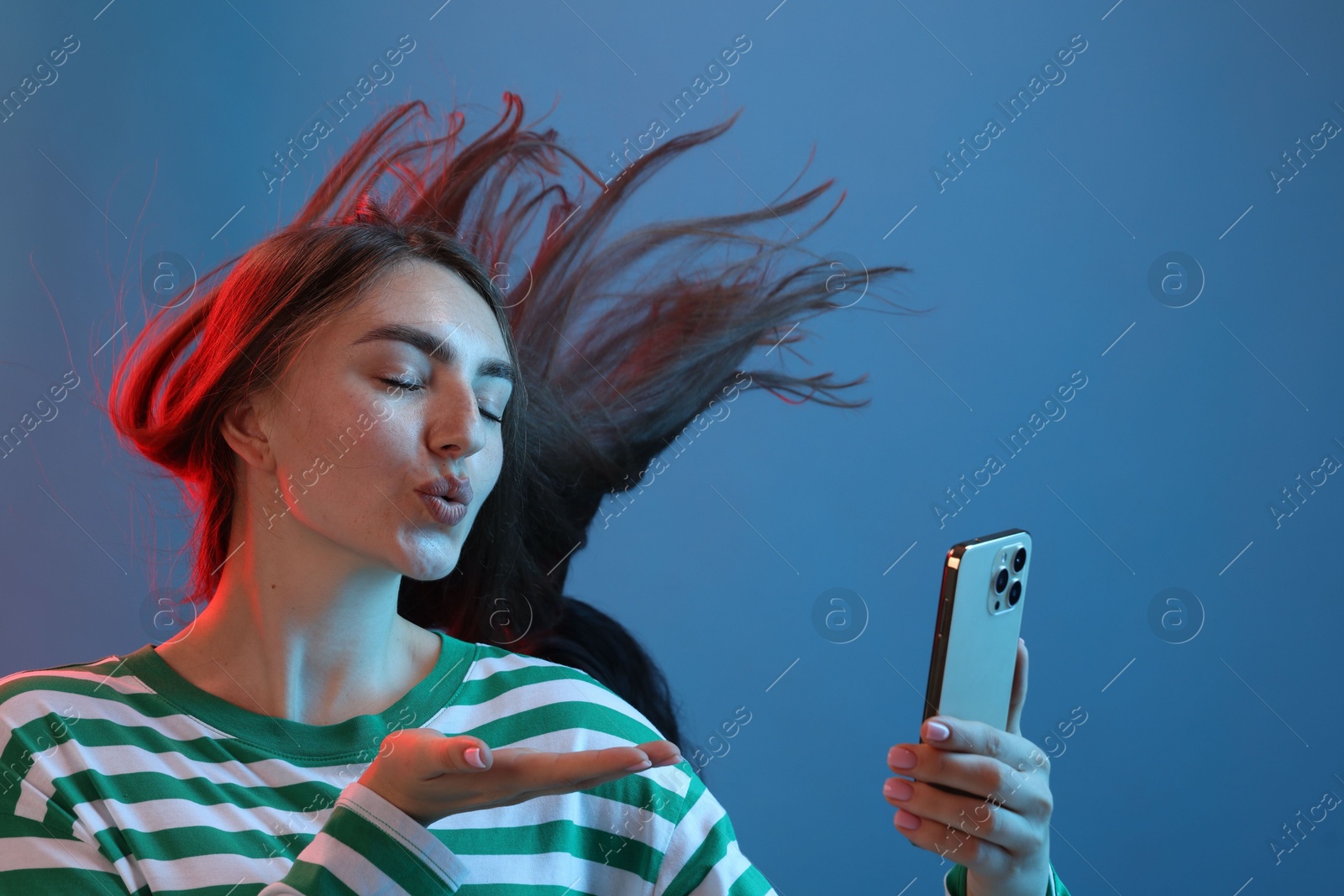 Photo of Young woman with beautiful long hair having videochat by smartphone and sending air kiss in neon lights against blue background
