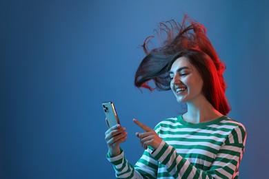 Photo of Happy woman with beautiful long hair using smartphone in neon lights against blue background. Space for text