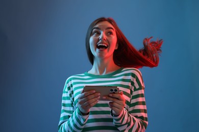 Photo of Happy woman with beautiful long hair using smartphone in neon lights against blue background, low angle view