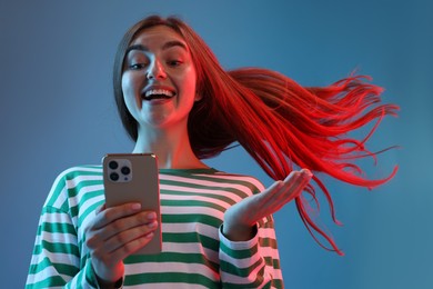 Photo of Happy woman with beautiful long hair using smartphone in neon lights against blue background, low angle view