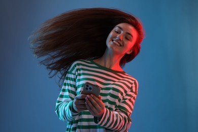 Photo of Happy woman with beautiful long hair using smartphone in neon lights against blue background, low angle view