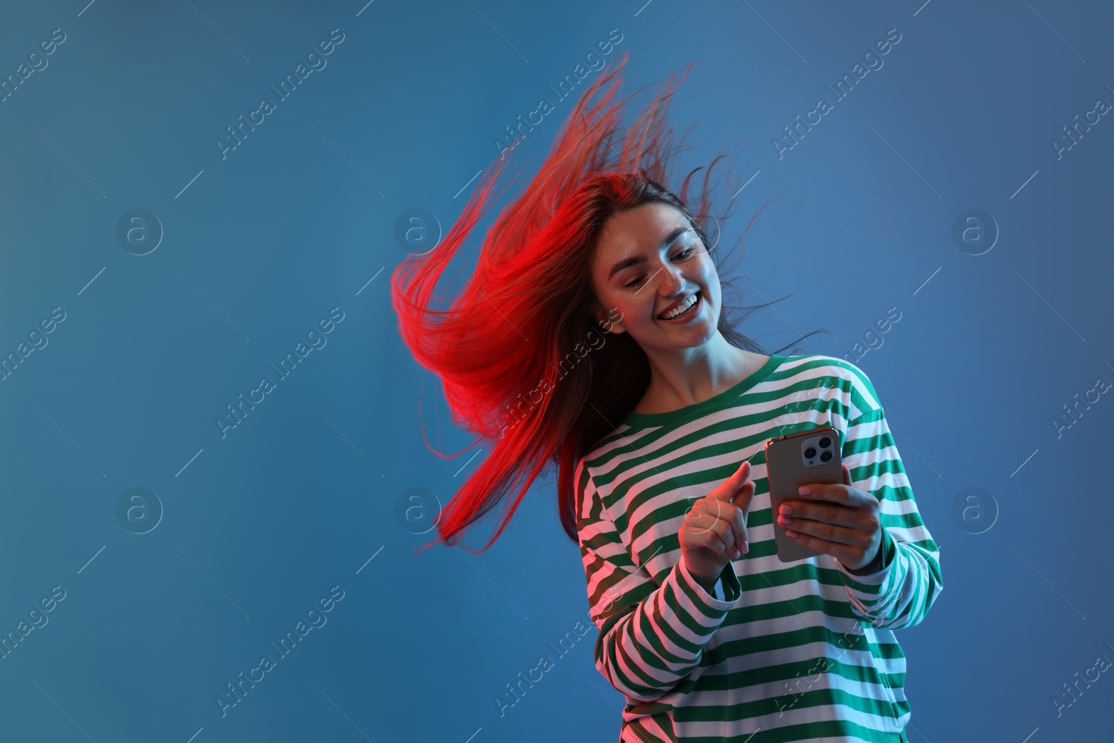 Photo of Happy woman with beautiful long hair using smartphone in neon lights against blue background. Space for text