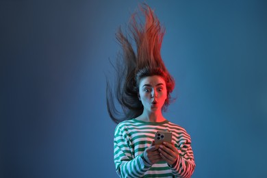 Photo of Surprised woman with beautiful long hair using smartphone in neon lights against blue background. Space for text