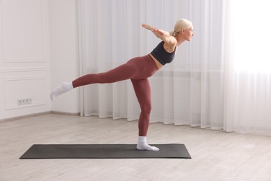 Photo of Mature woman in sportswear stretching on mat near window at home