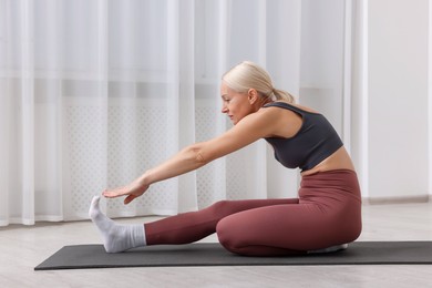 Photo of Mature woman in sportswear stretching on mat near window at home