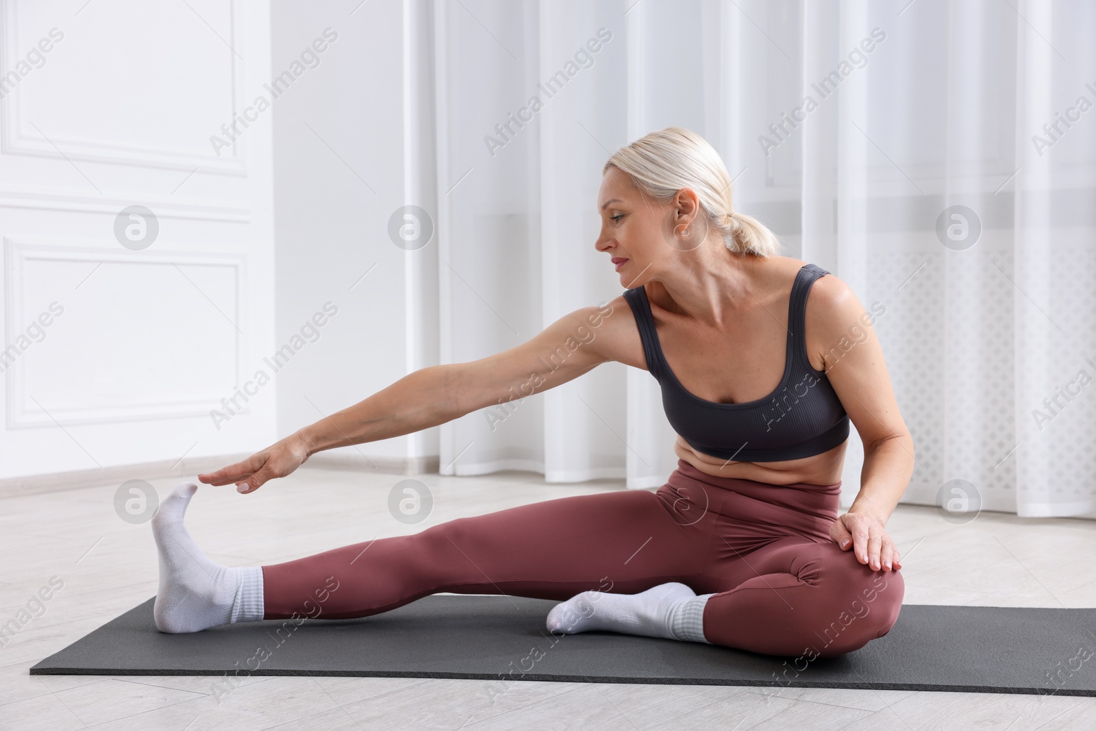 Photo of Mature woman in sportswear stretching on mat near window at home