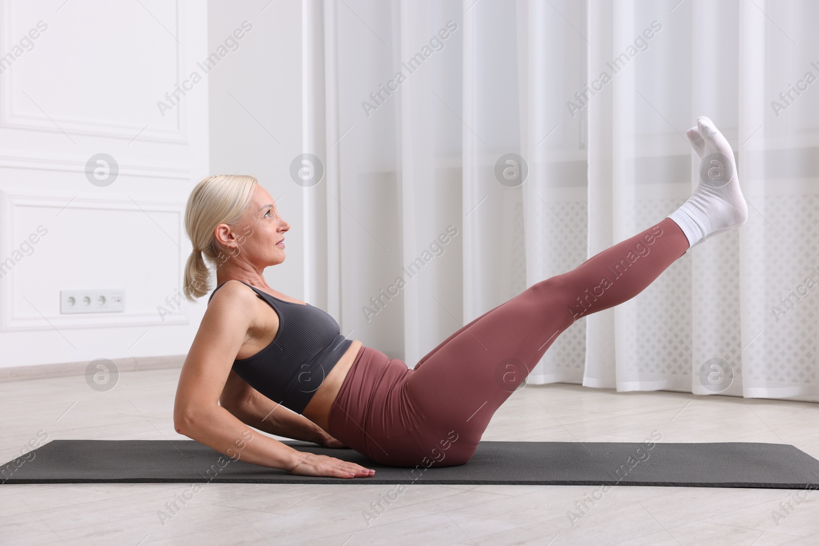 Photo of Mature woman in sportswear stretching on mat near window at home