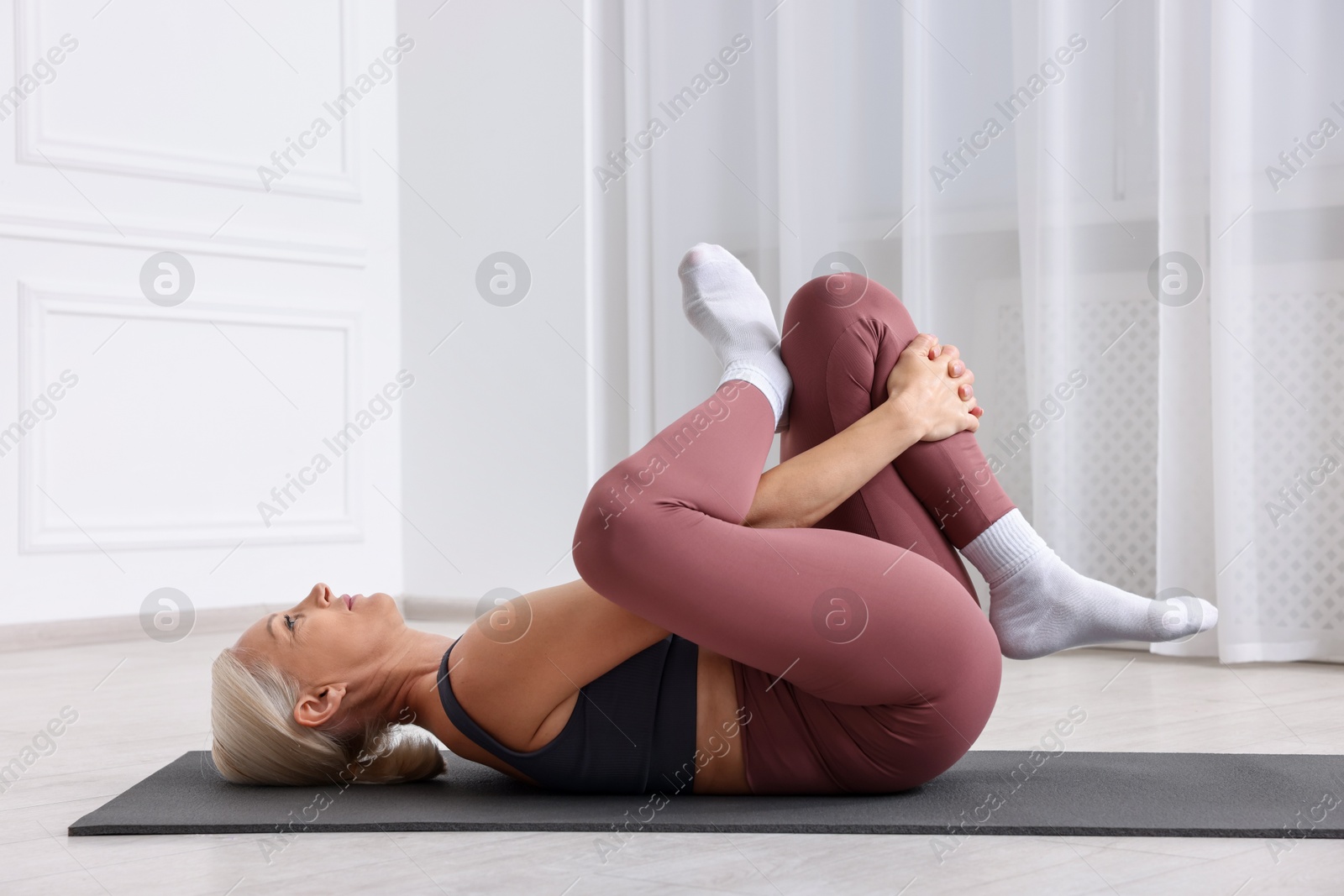 Photo of Mature woman in sportswear stretching on mat near window at home