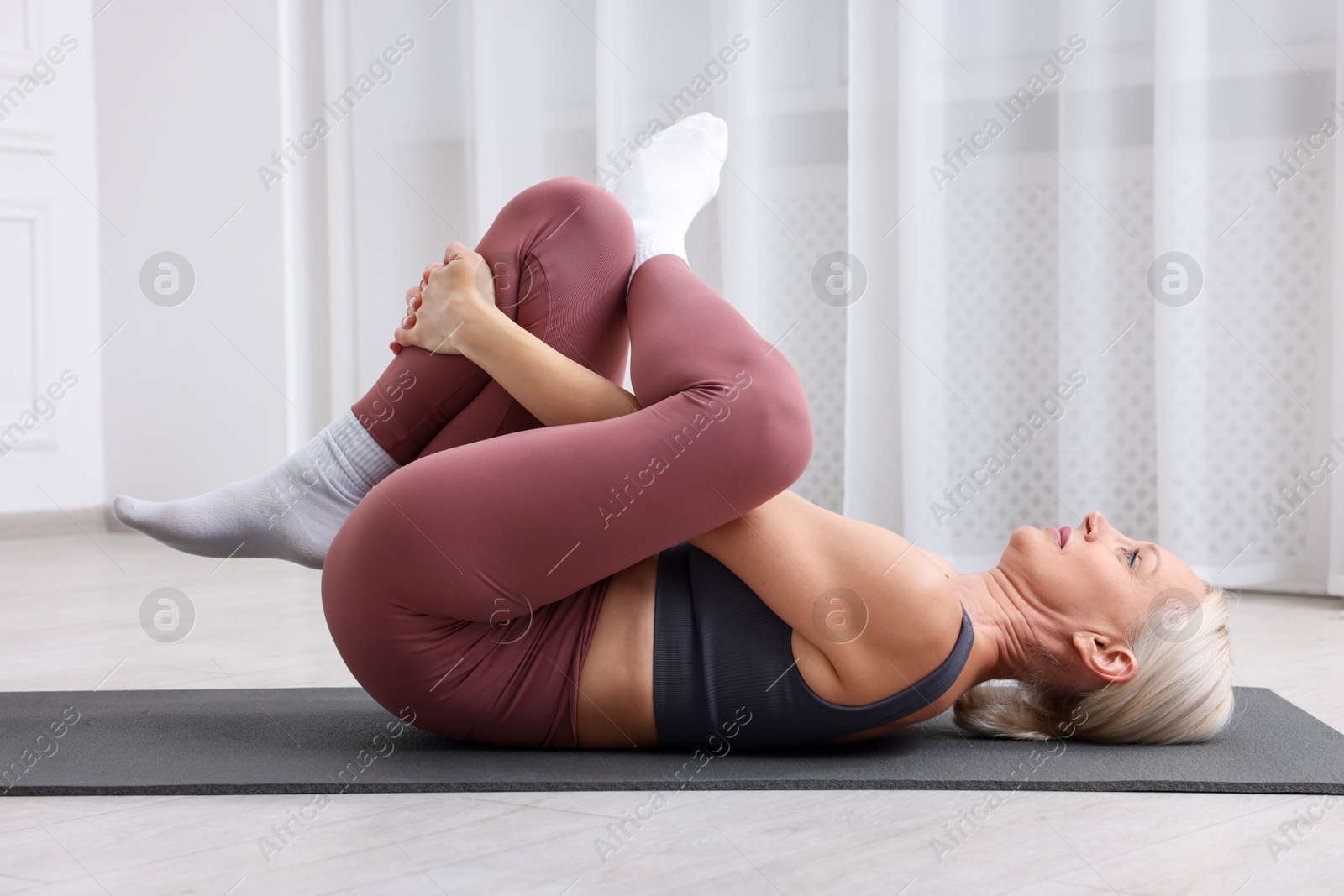 Photo of Mature woman in sportswear stretching on mat near window at home