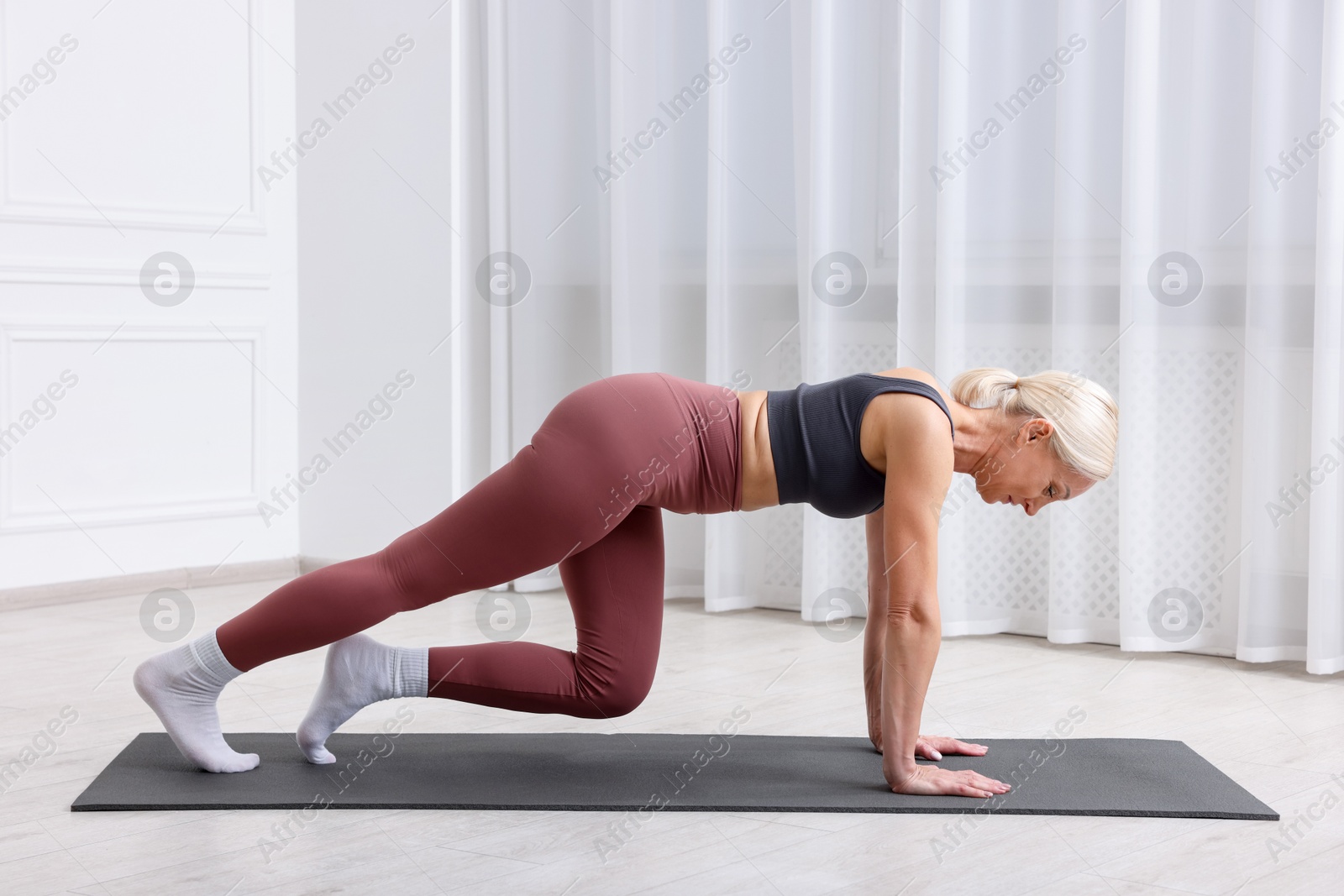 Photo of Mature woman in sportswear stretching on mat near window at home
