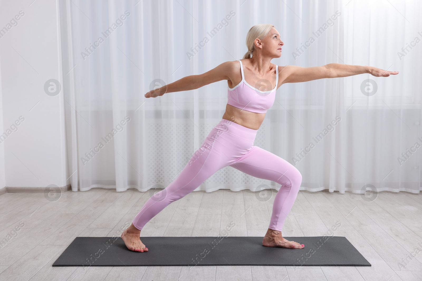 Photo of Mature woman in sportswear stretching on mat near window at home