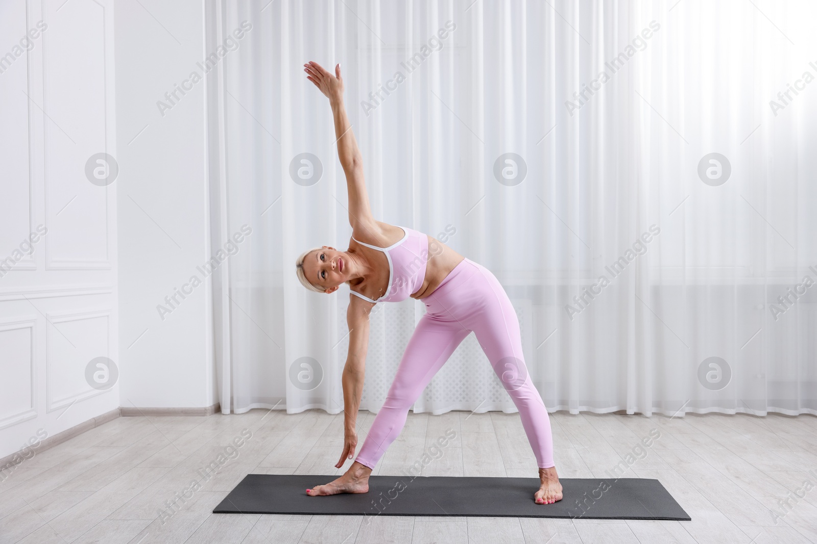 Photo of Mature woman in sportswear stretching on mat near window at home