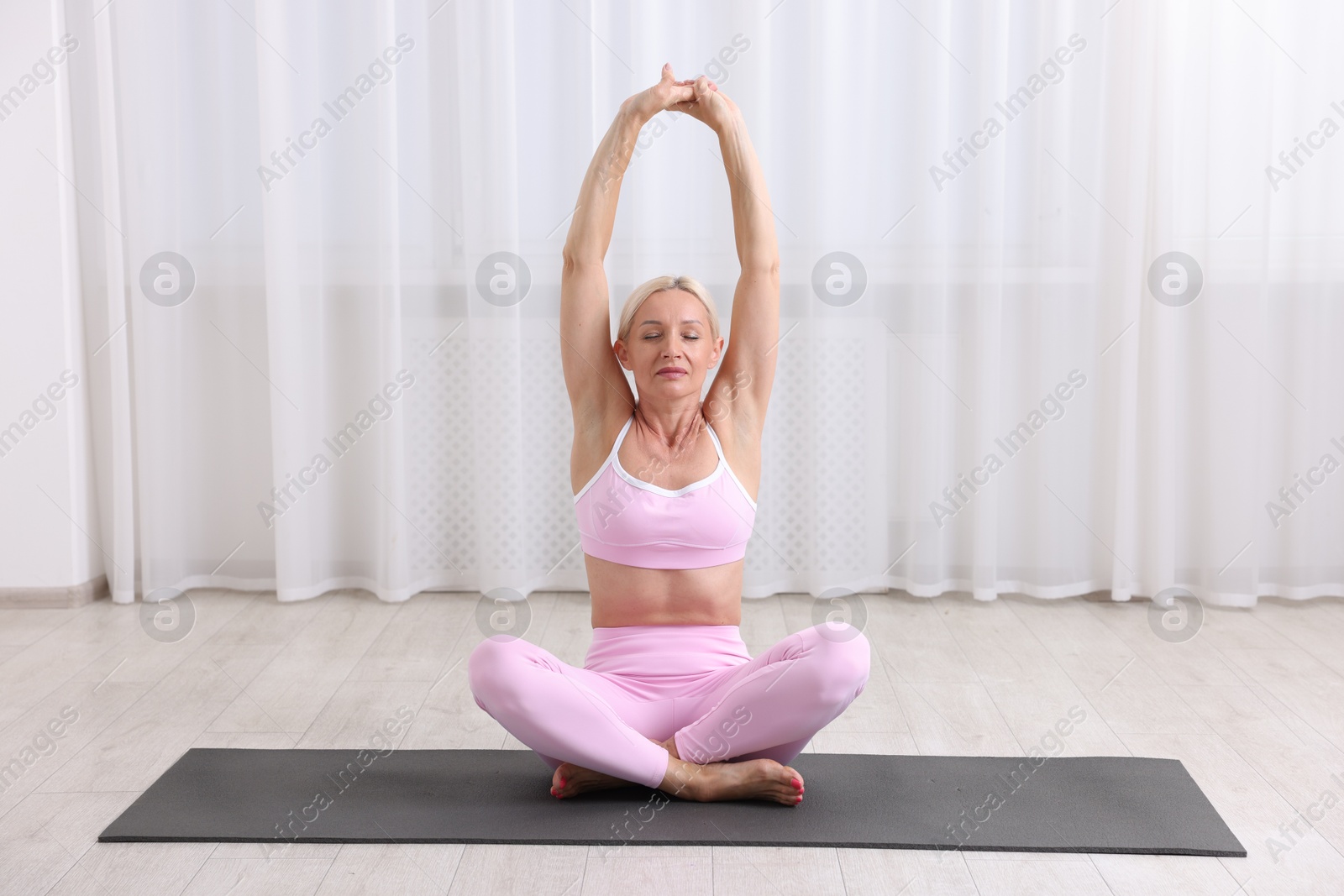 Photo of Mature woman in sportswear stretching on mat near window at home