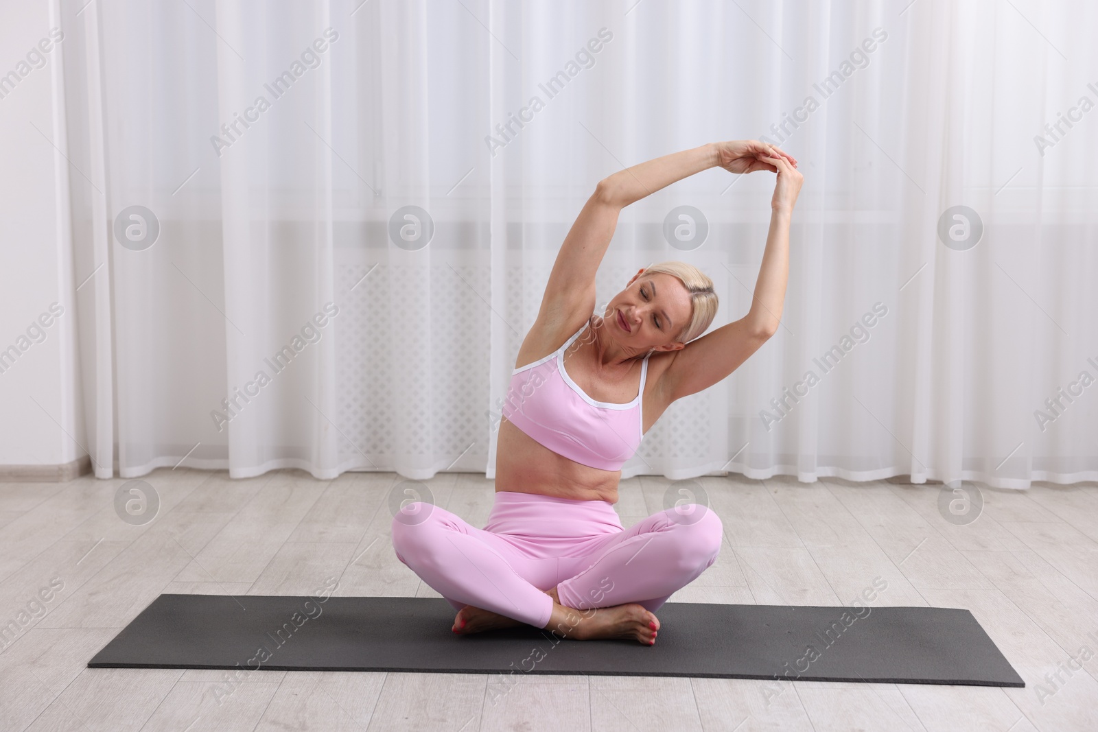 Photo of Mature woman in sportswear stretching on mat near window at home