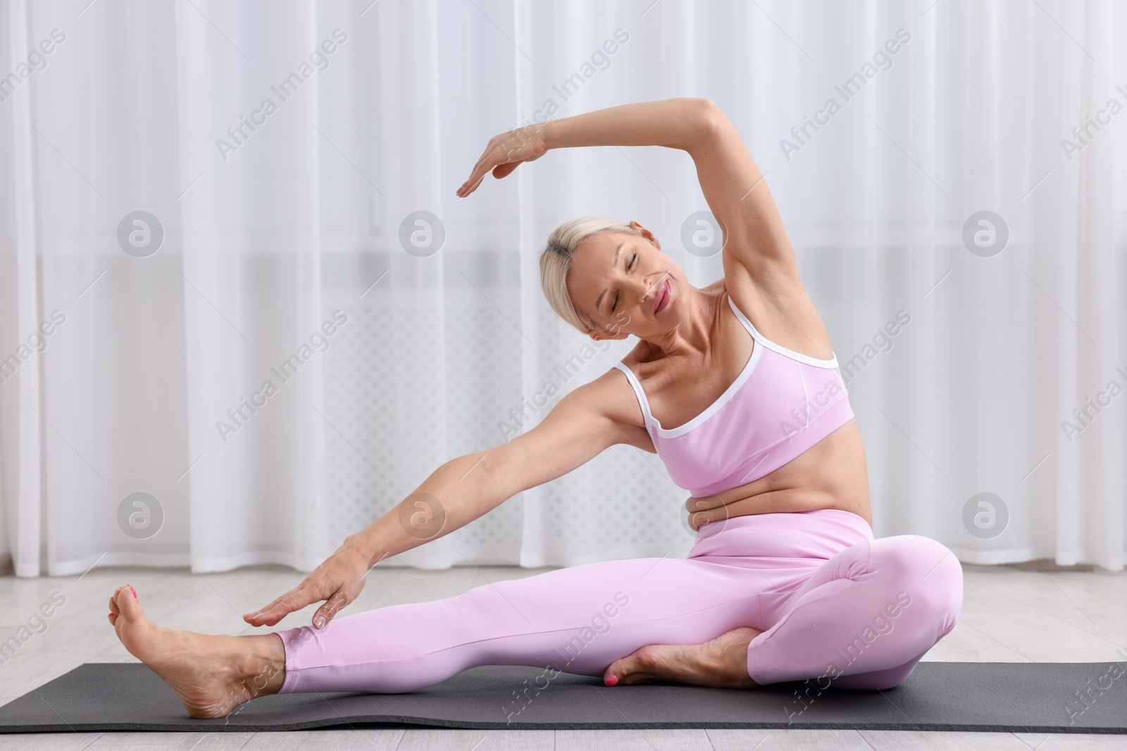 Photo of Mature woman in sportswear stretching on mat near window at home