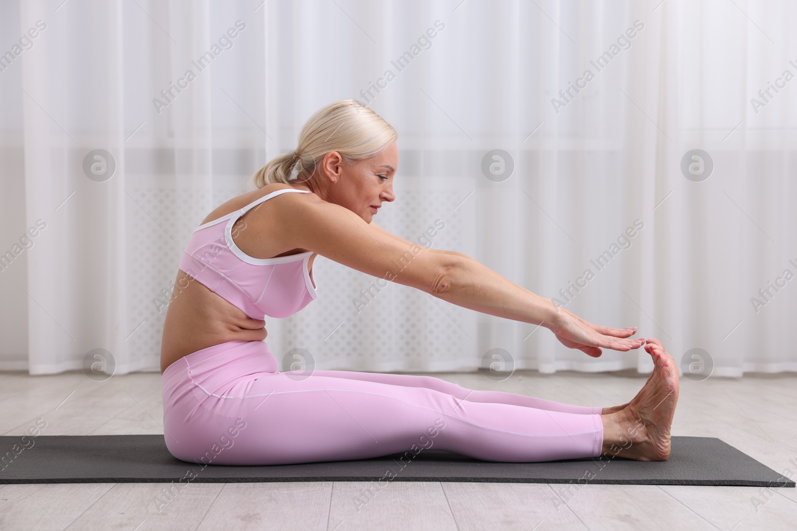 Photo of Mature woman in sportswear stretching on mat near window at home