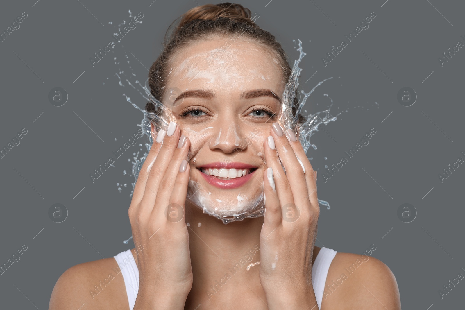 Young woman washing face with cleansing foam and water splashes on grey background Image of Young woman washing face with cleansing foam and water splashes on grey background