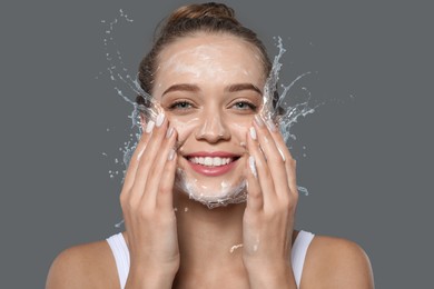 Image of Young woman washing face with cleansing foam and water splashes on grey background
