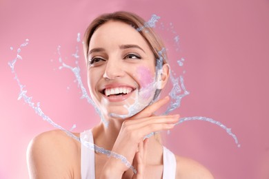 Image of Young woman washing face with scrub and water splashes on pink background