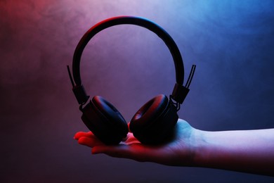 Photo of Woman holding wireless headphones on dark background with color lights and smoke, closeup