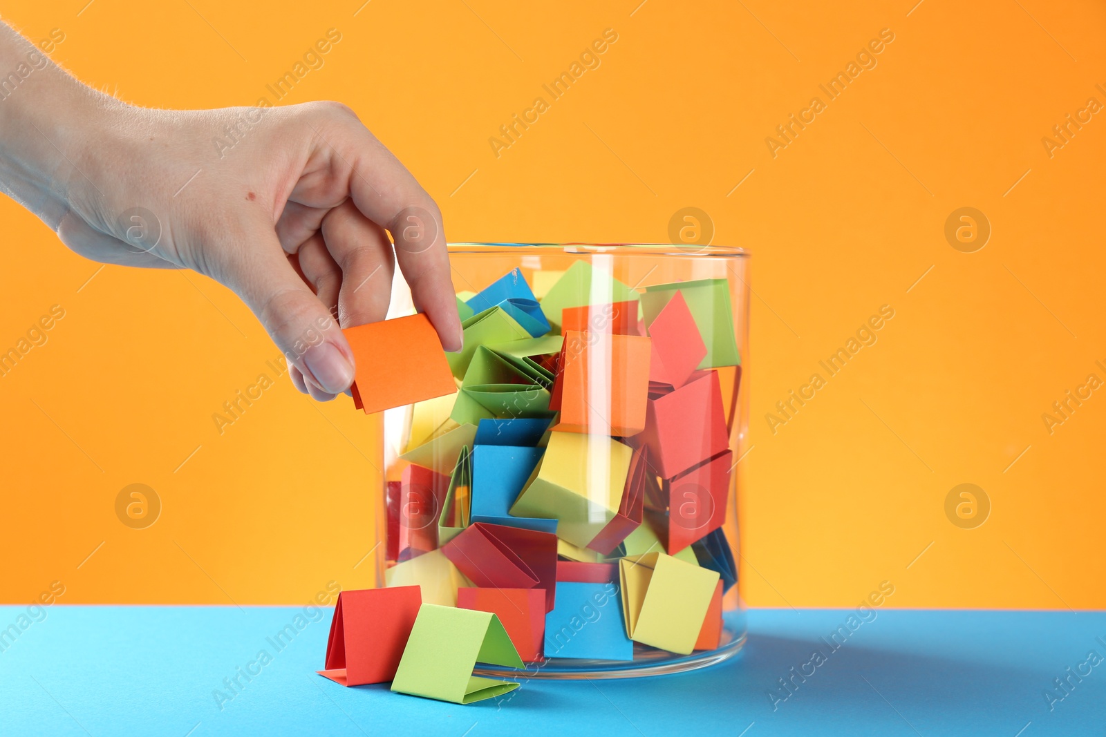 Woman taking orange paper piece from glass jar on color background, closeup Photo of Woman taking orange paper piece from glass jar on color background, closeup