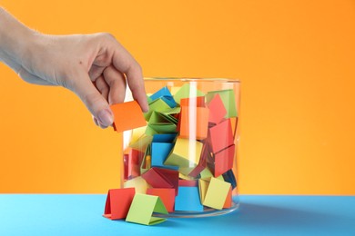 Photo of Woman taking orange paper piece from glass jar on color background, closeup