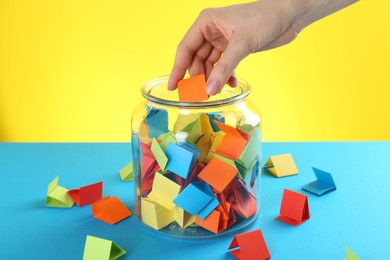 Photo of Woman taking orange paper piece from glass jar on color background, closeup