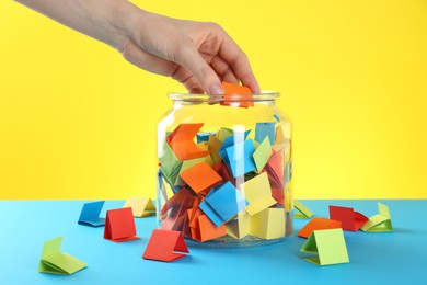 Photo of Woman taking orange paper piece from glass jar on color background, closeup