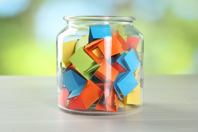 Photo of Colorful paper pieces in glass jar on wooden table against blurred background, closeup