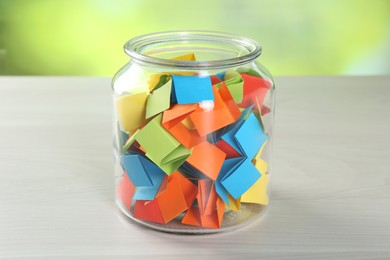 Photo of Colorful paper pieces in glass jar on wooden table against blurred background, closeup